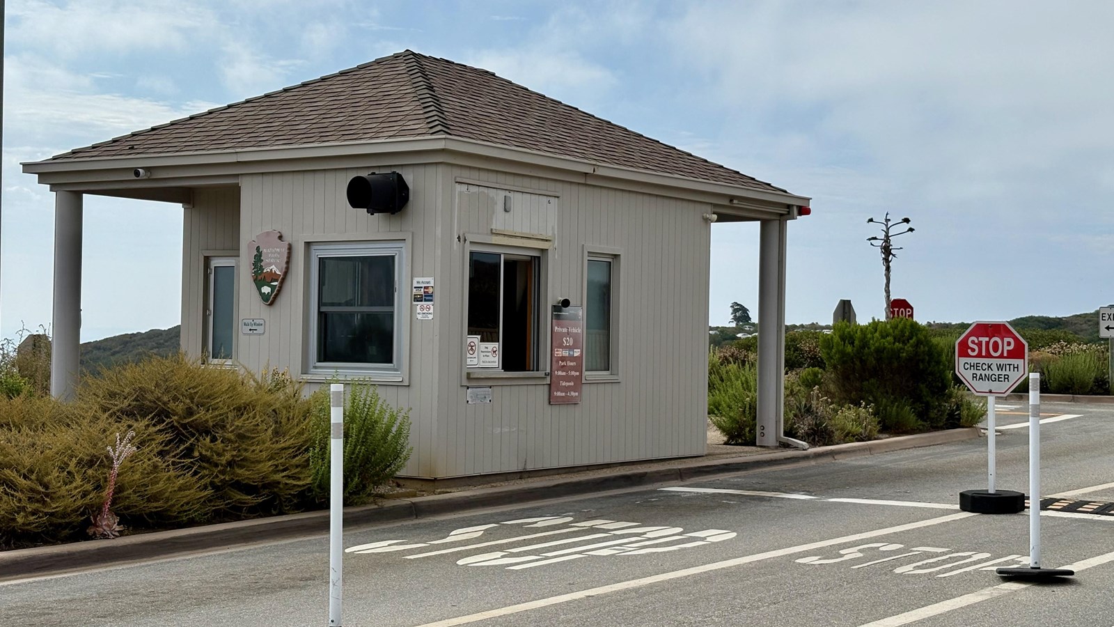 Small ranger station with stop signs, entry fee sign, and bushes under a partly cloudy sky.