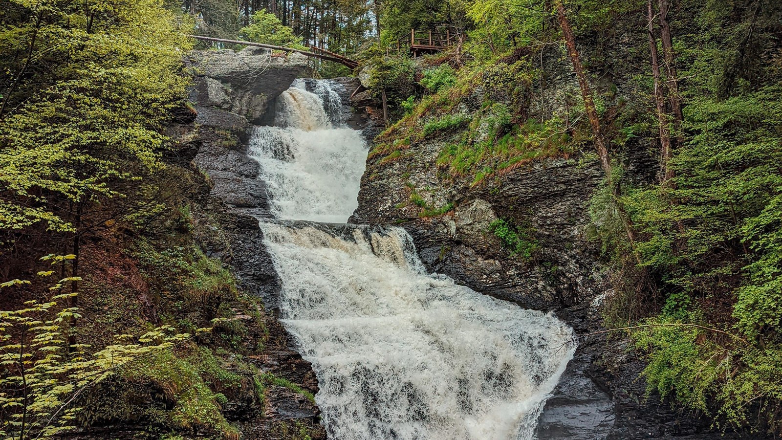 A tiered waterfall surrounded by rock cliffs and green foliage.