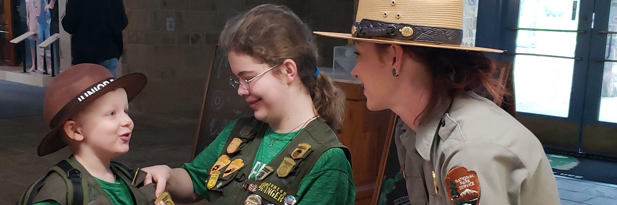 A park ranger and two junior rangers at the front desk with junior ranger badges.