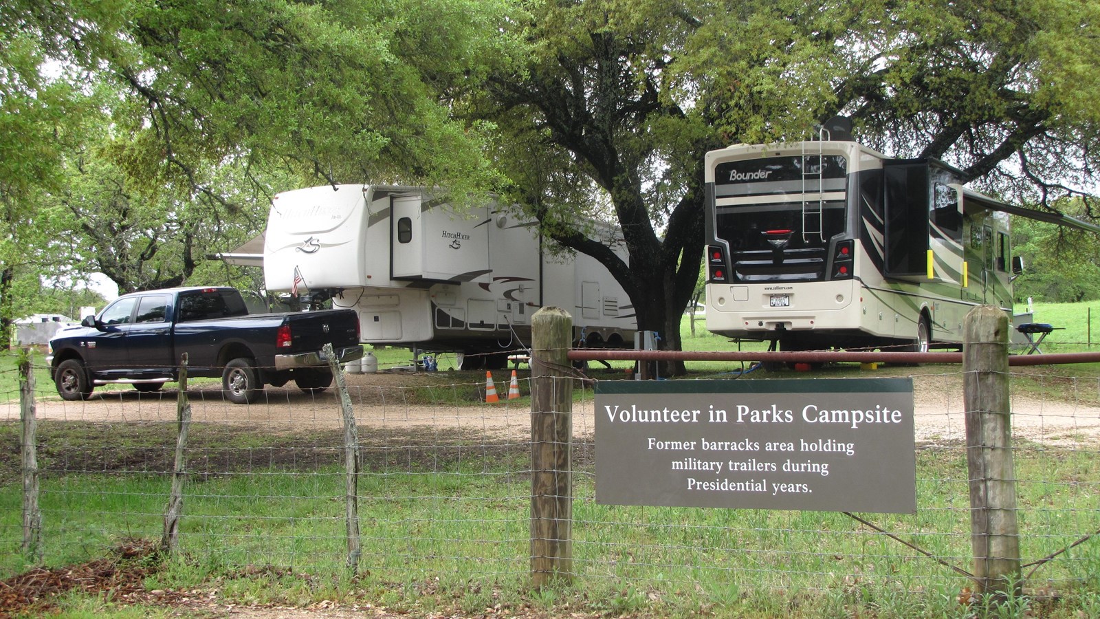 Two RVs sit under the shade of trees within an enclosed Volunteers-In-Parks campsite area.