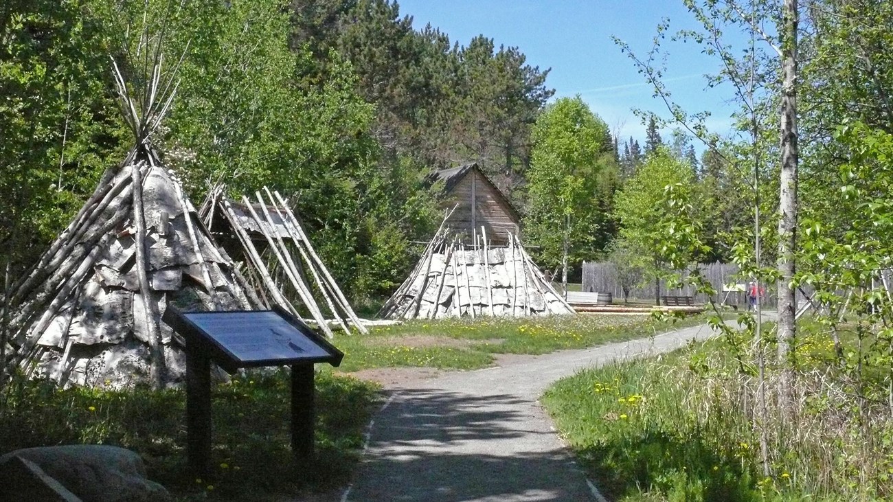 Bark dwellings along a gravel path in front of a reconstructed historic wooden building.