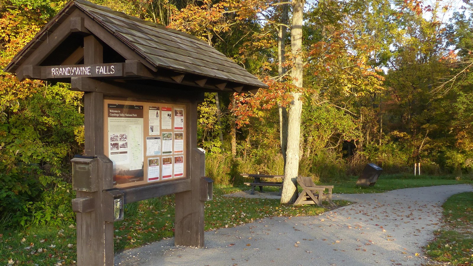 A roofed “Brandywine Falls” information kiosk stands along a paved path leading past picnic tables.