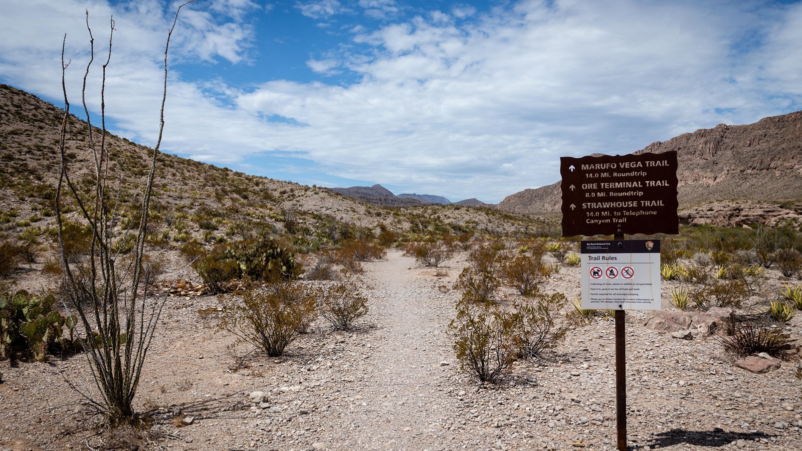 A metal sign with trail information sits beside a dirt path heading into the mountains.