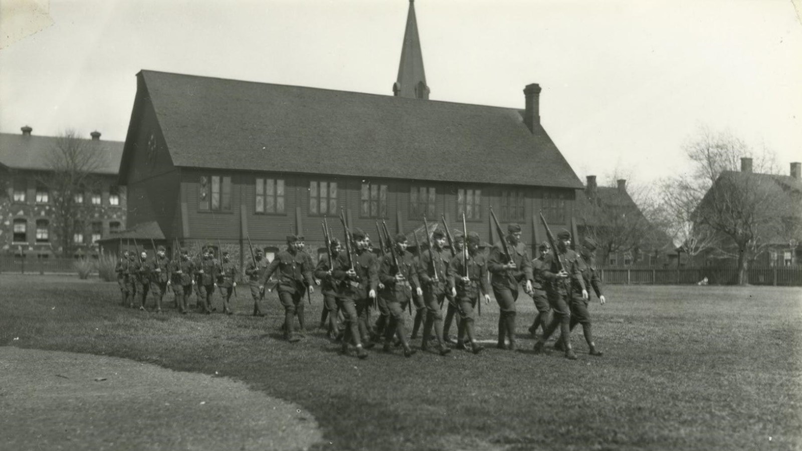 Black and white historic image of men with rifles walking on a grassy field with a church behind.