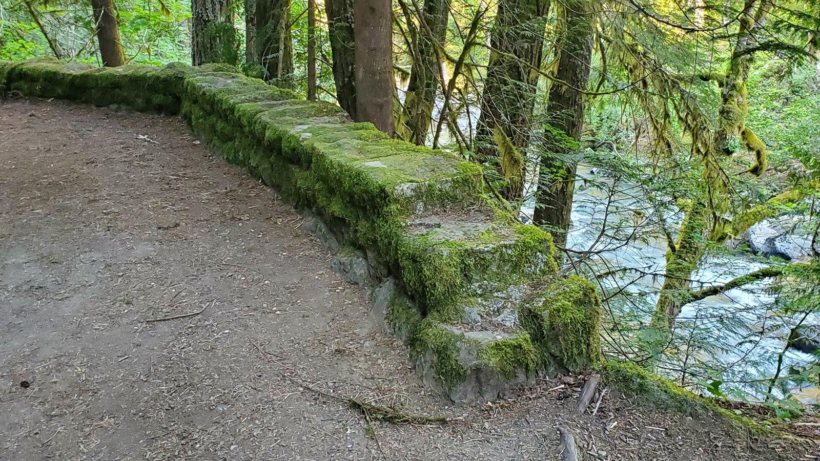 2 foot tall rock wall between the trail and a steep forested slope. River in the background.