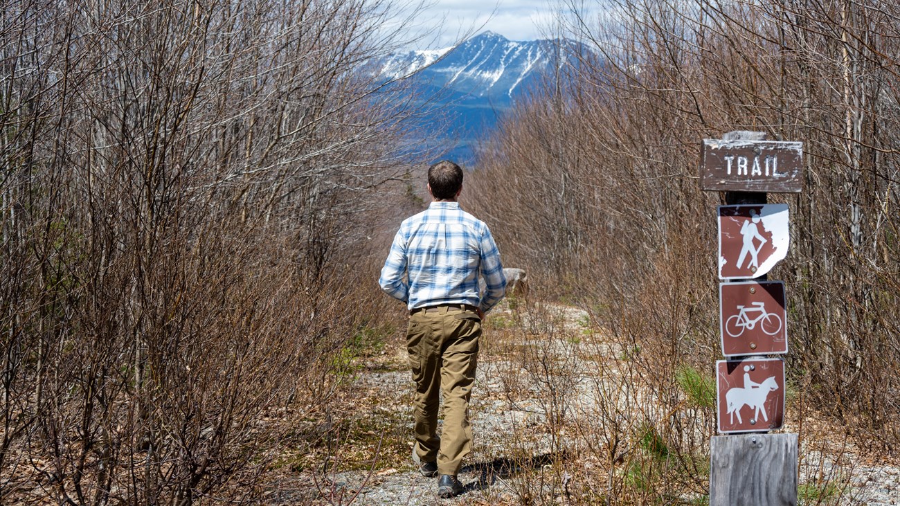 A hiker at the gravel trailhead area with a view of Katahdin, a tall mountain in the distance.