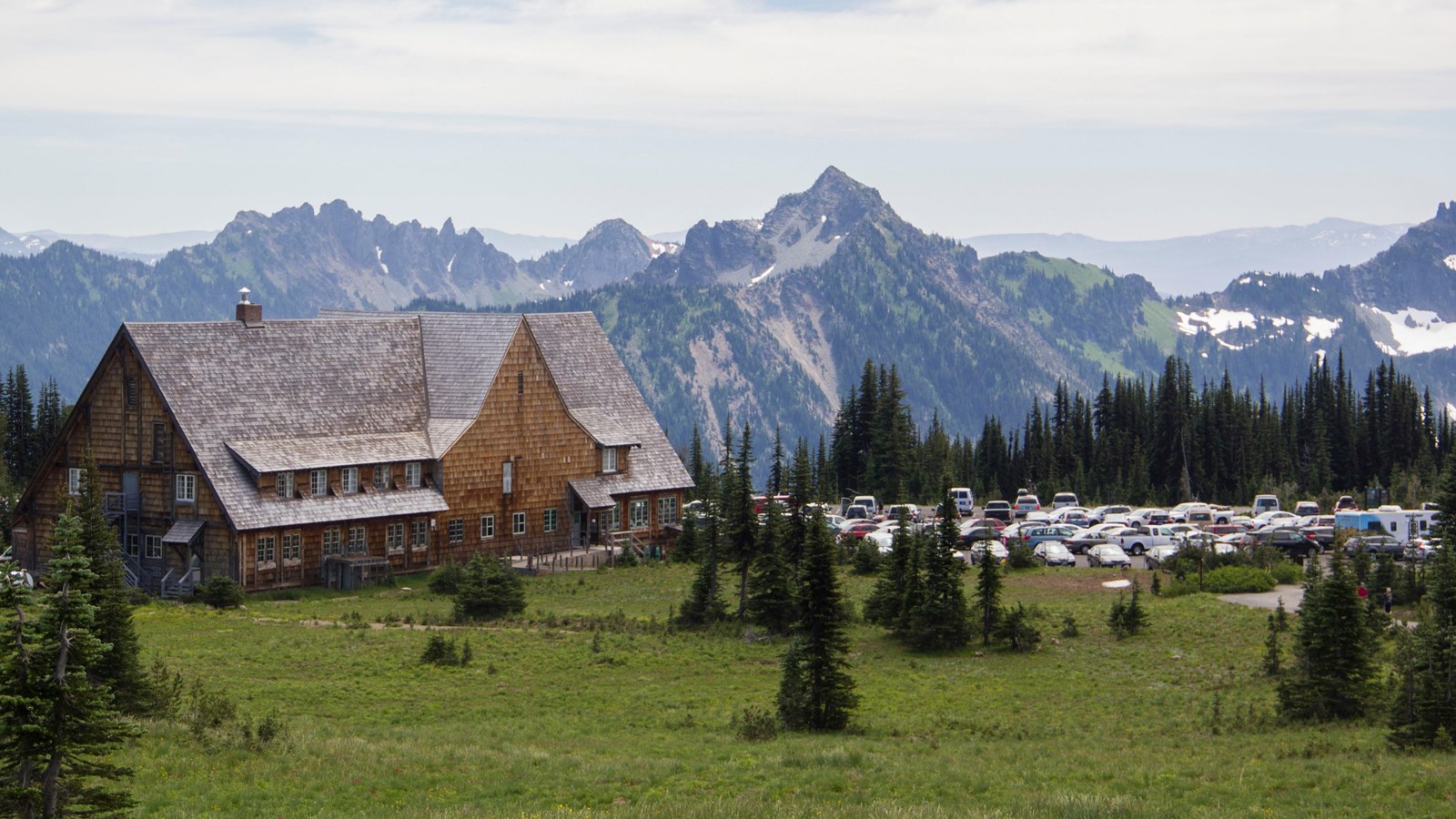 A large wood building with a shingle roof nestled in a mountain meadow next to a parking area.
