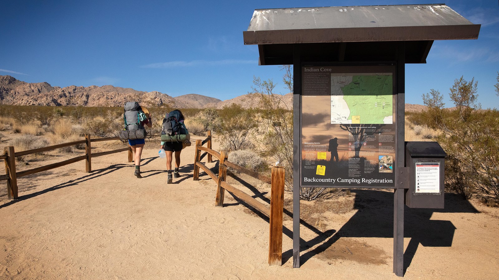 Two people walking down a dirt trail between two split rail fences past a bulletin board.