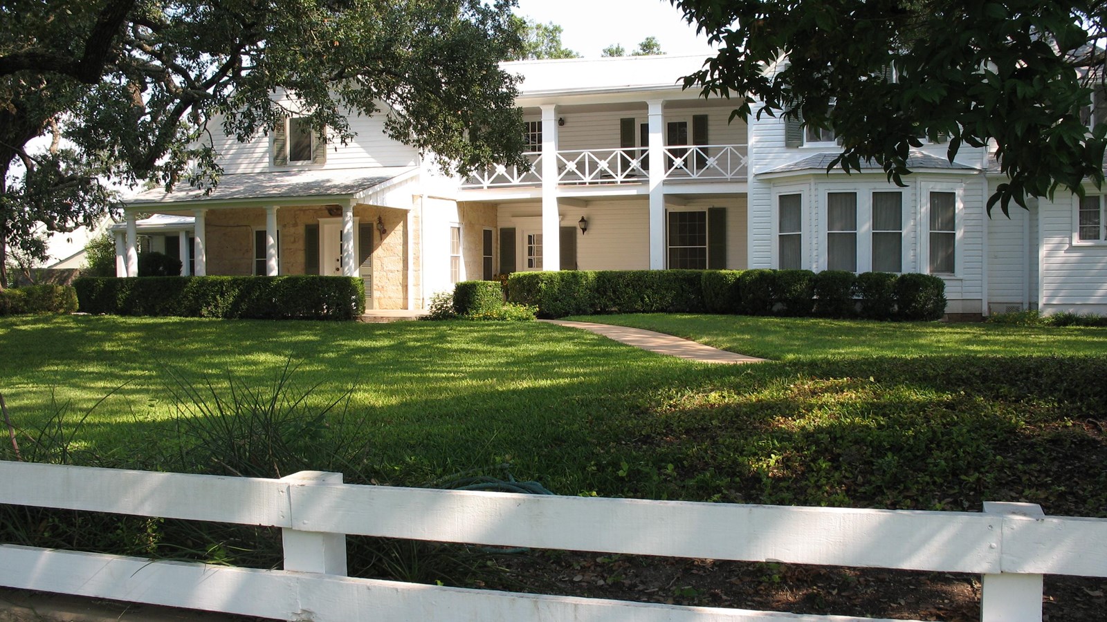 A large, white, two-story home is shaded by massive oak trees.