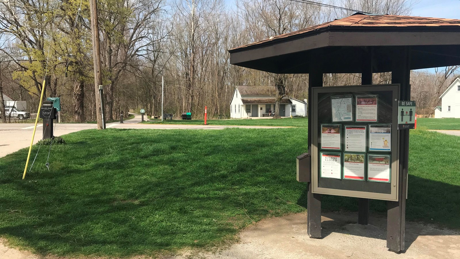 Information kiosk stands to the right of a gray trail that crosses a road to a small white house.