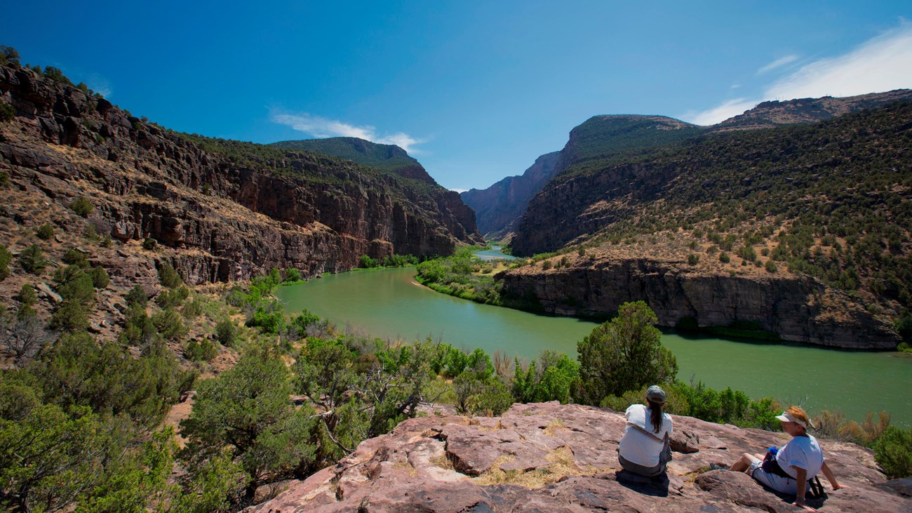 Two women sit on a large rock as a green colored river flows into a canyon in the distance.