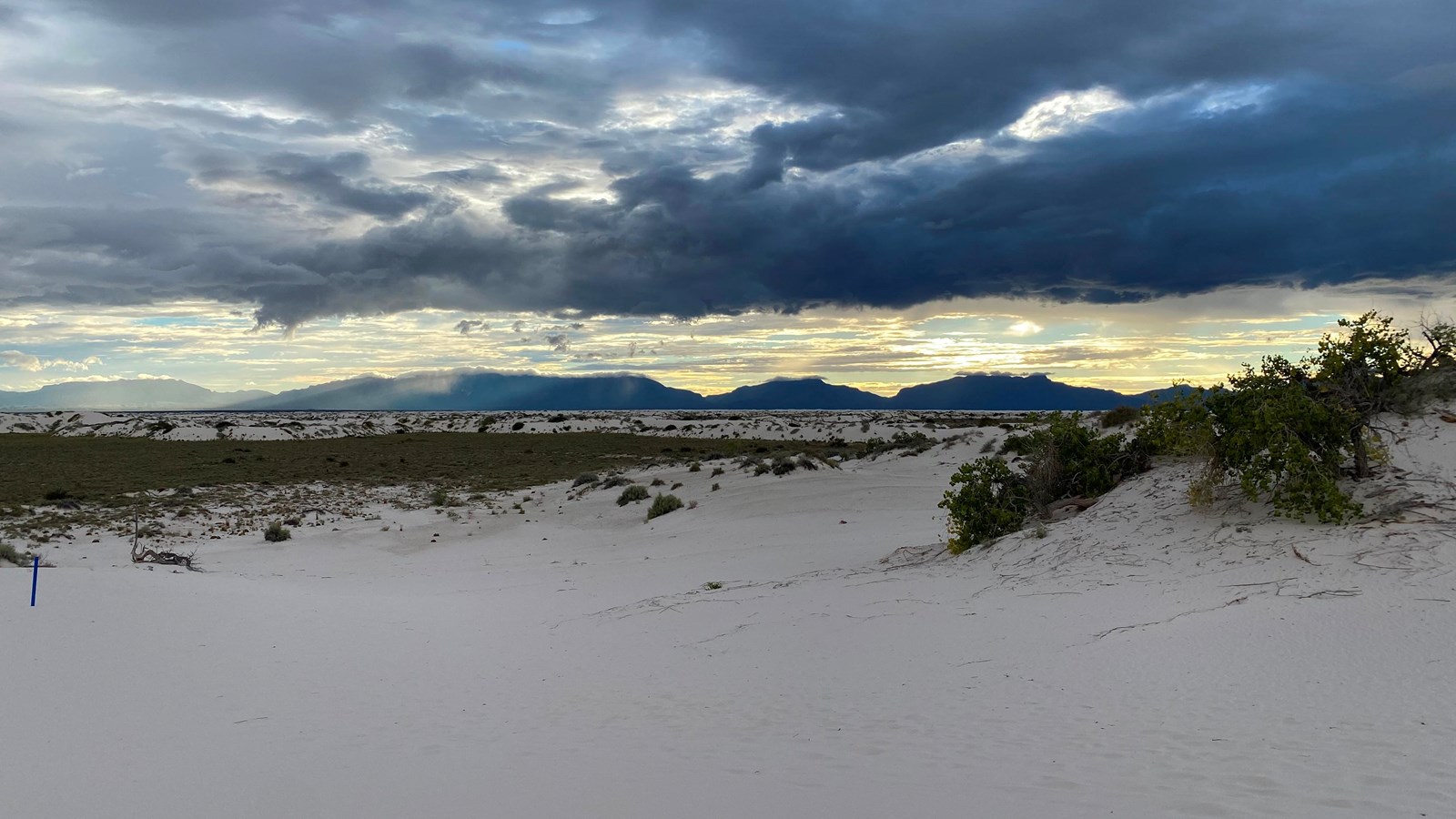 A white dune slopes toward the vegetation of the desert floor with a mountain range in the distance.