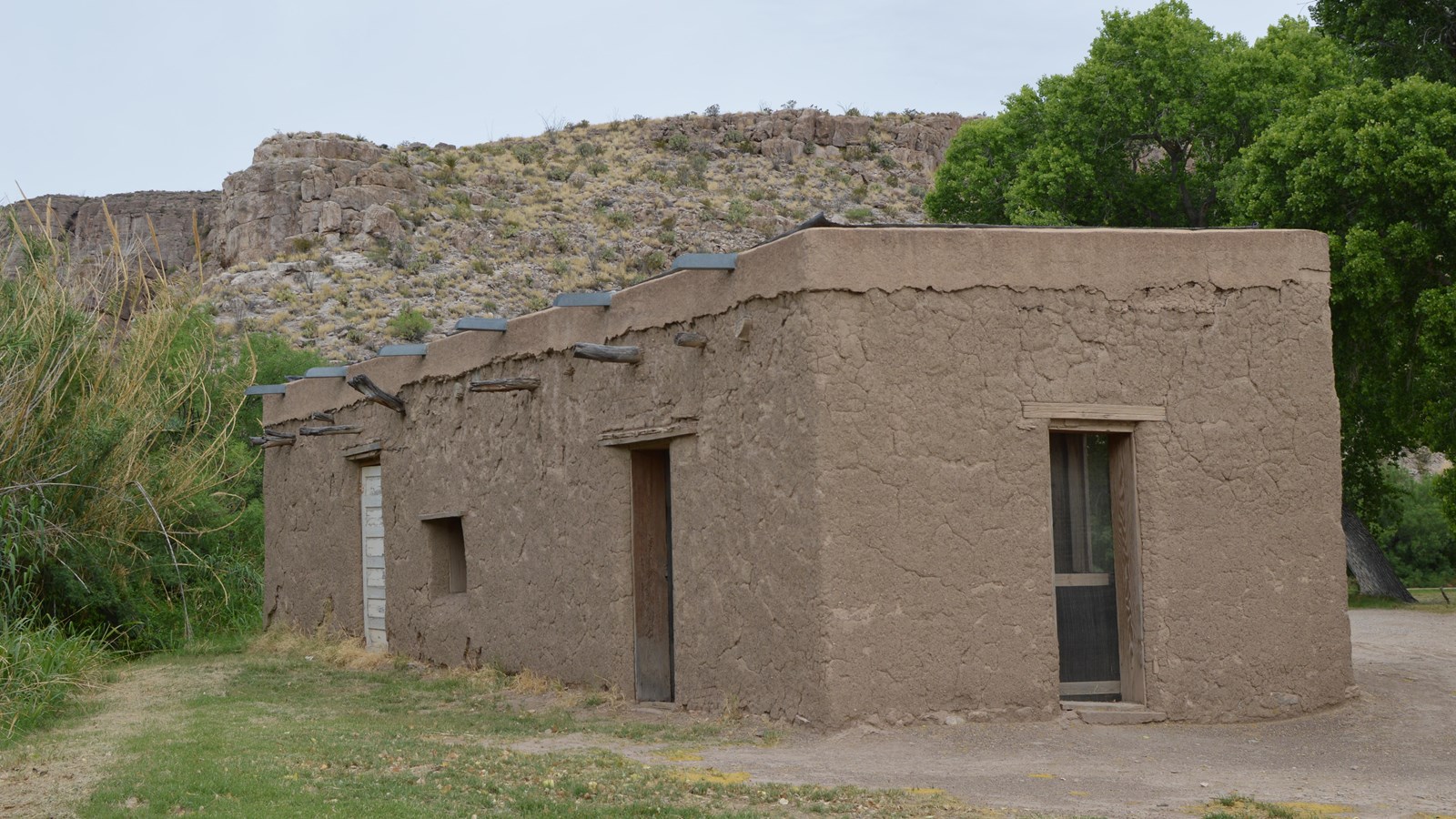 An small adobe building sits in a flat near cottonwood trees and limestone cliffs.