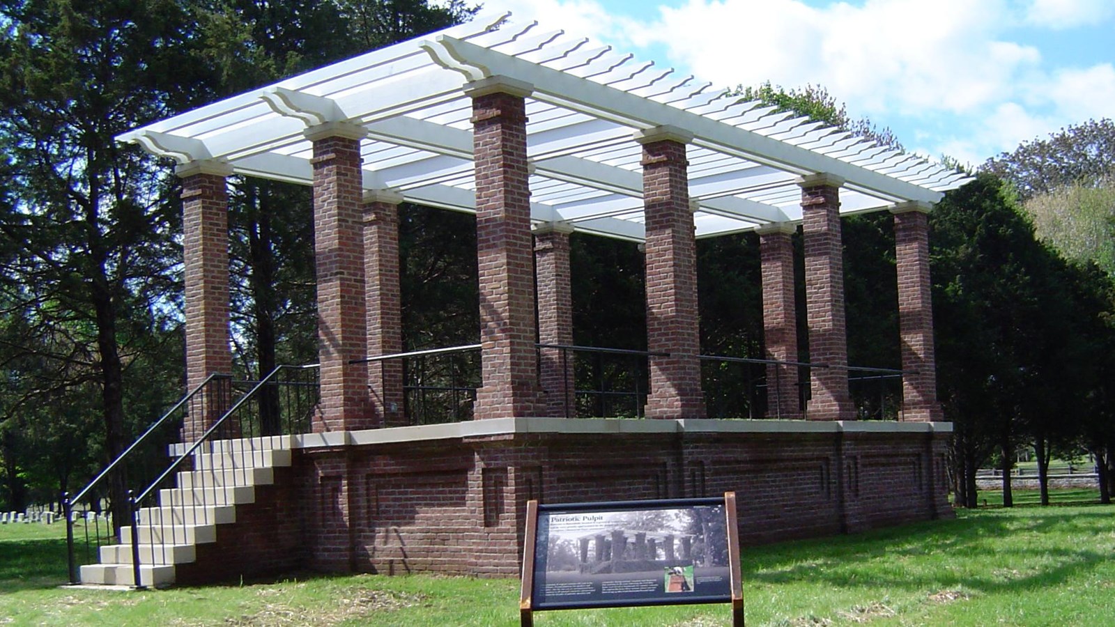 A brick platform with brick pillars supporting an open roof of white wood slats.