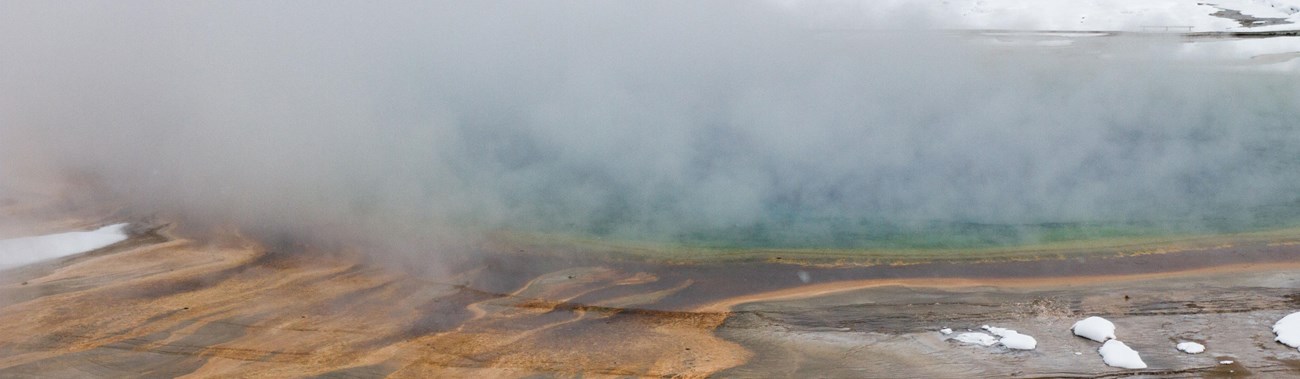 Steam rises from the prismatic pool with snow surrounding it.
