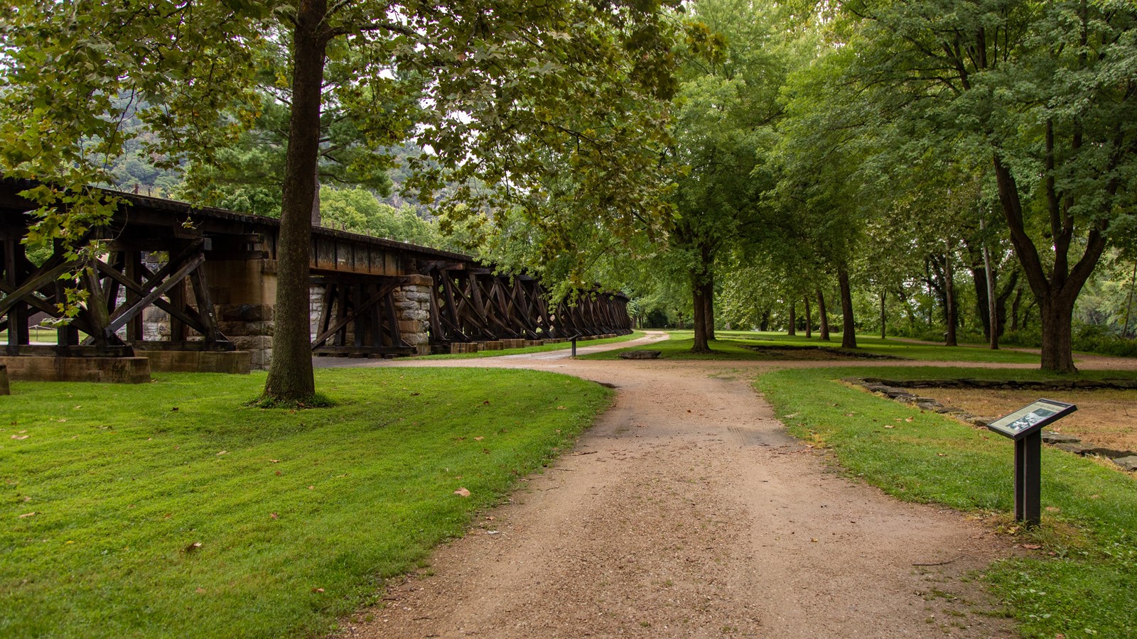 A gravel road curves under a train trestle.