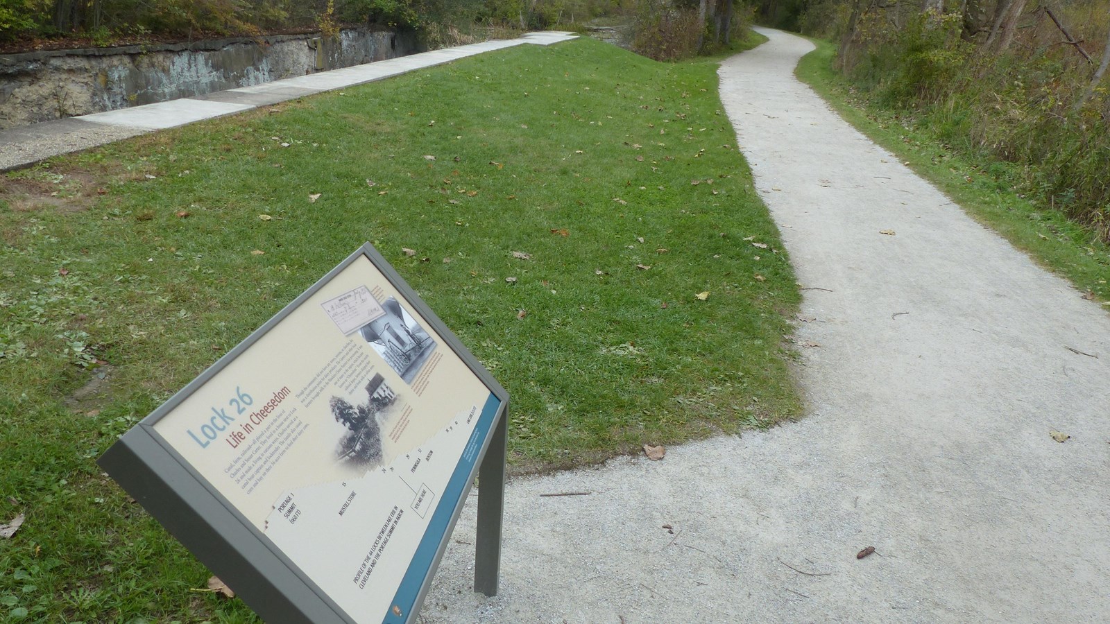 A pale gray trail to the right of and parallel to a stone canal lock and graphic panel.