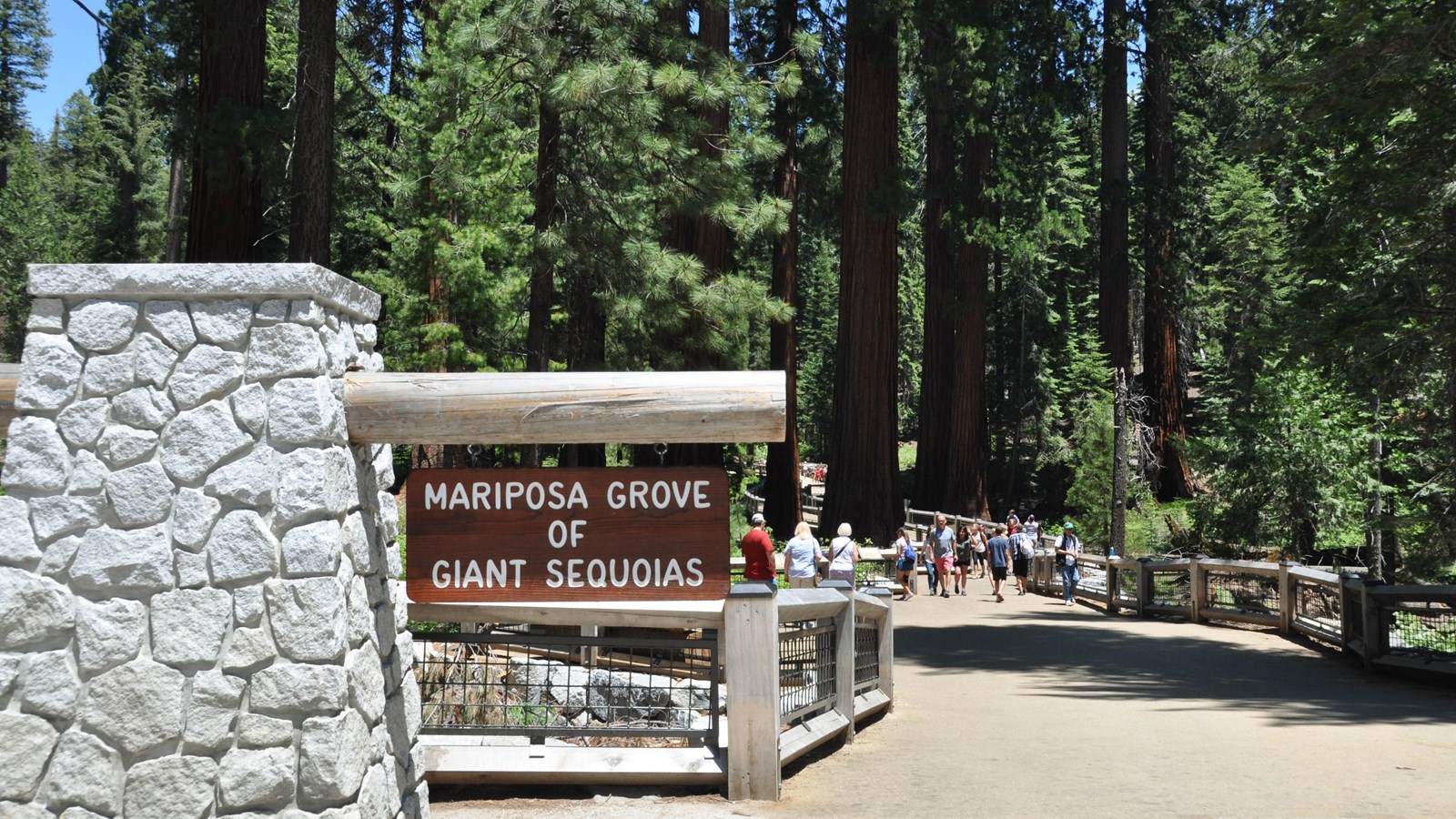 Wide flat accessible trail with place name sign and people walking on fence lined trail