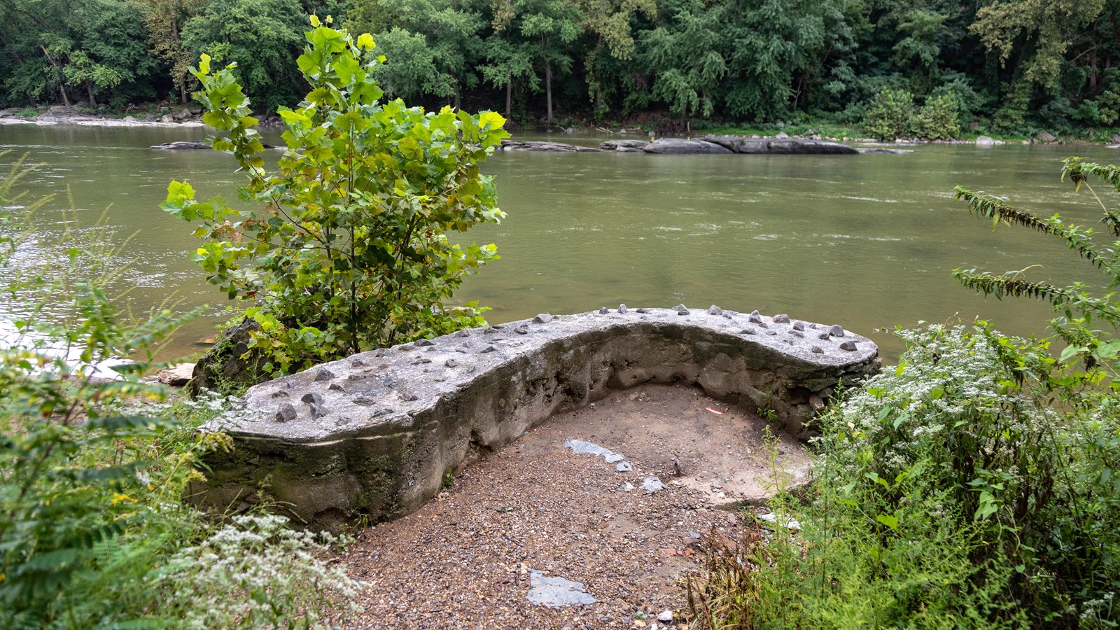 A curved stone and cement foundation with jagged rocks on top adjacent to the river.