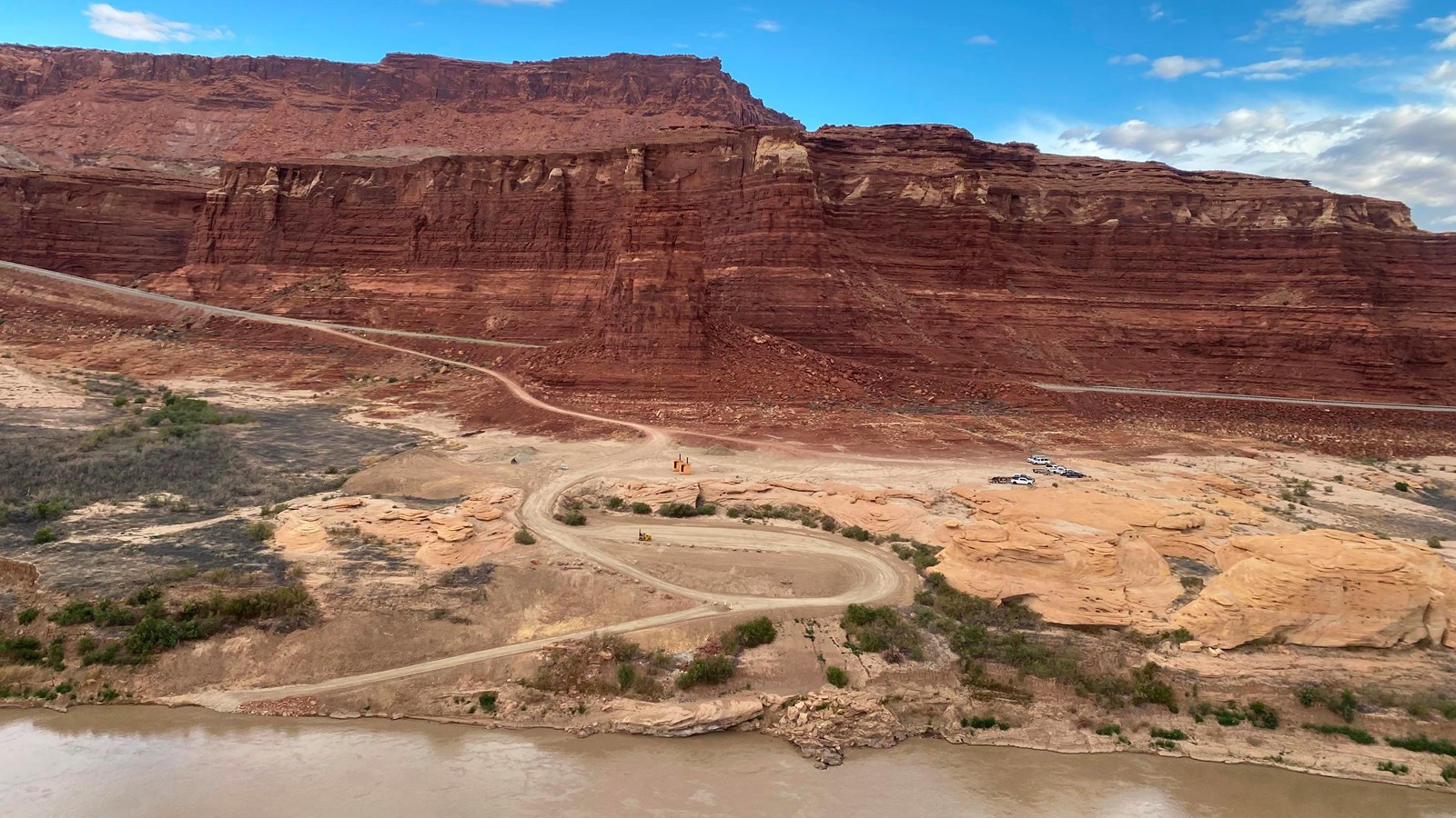 An unpaved boat takeout along muddy water, under red rock cliffs and blue skies.