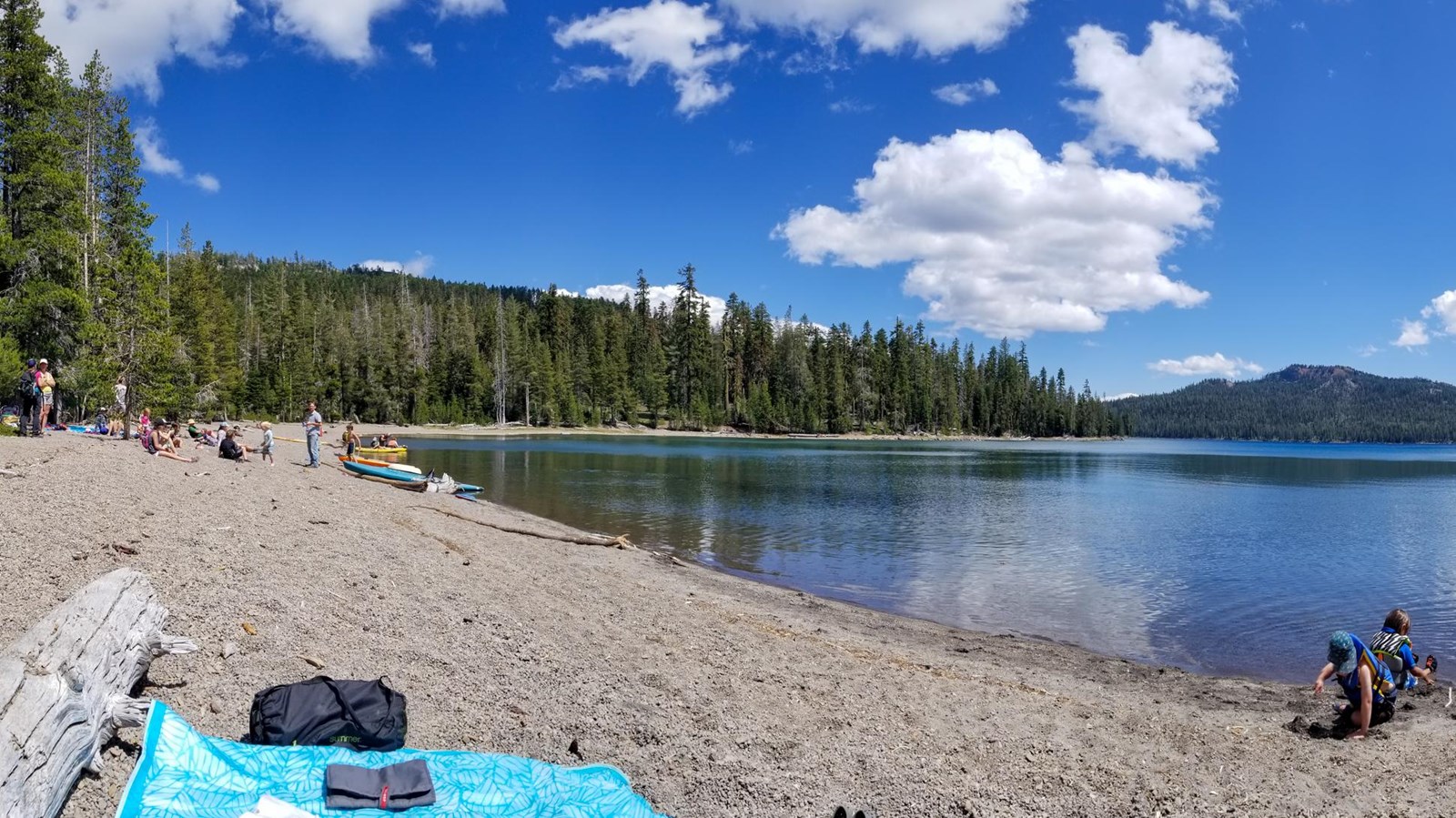 People on a gravel beach on the shore of a mountain lake lined by conifers and a large volcanic peak