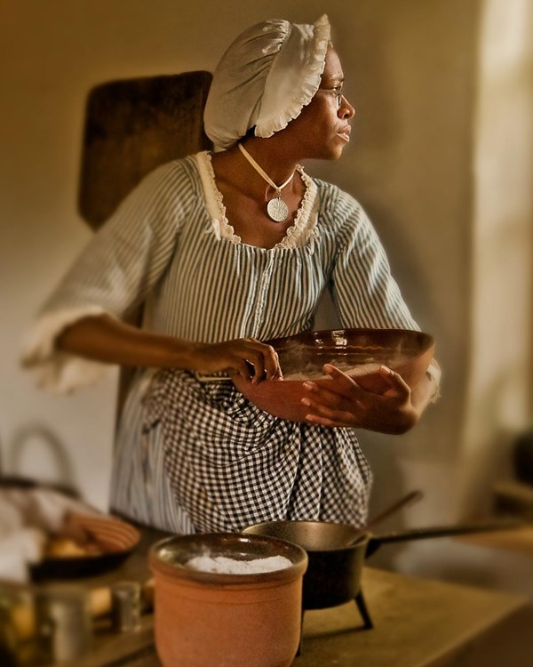 A dark-skinned woman holding a mixing bowl looks out a window