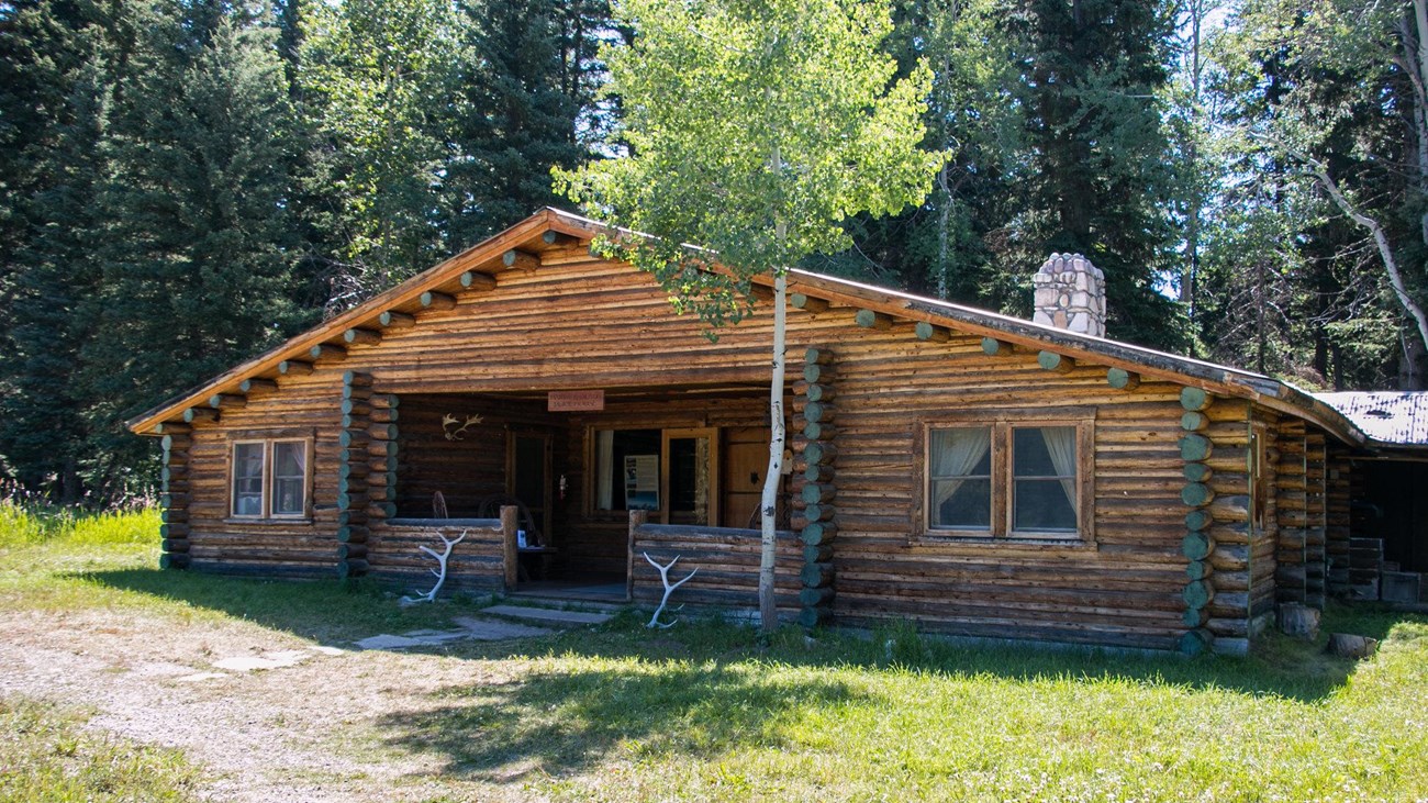 A brown, log cabin building with a river stone chimney and elk antlers resting on the front porch.