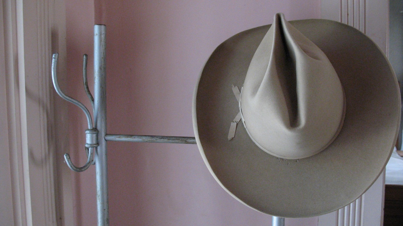 A tan colored cowboy hat hangs on a silver coat tree. Pink wallpaper covers the wall behind it.