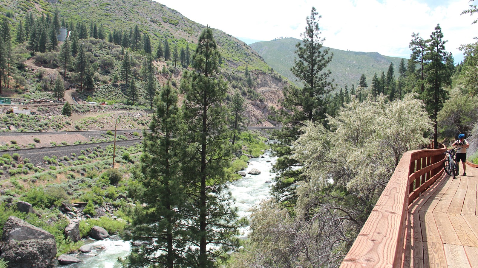 Boardwalk with a cyclist on the right, rushing whitewater and forested mountainside on the left.