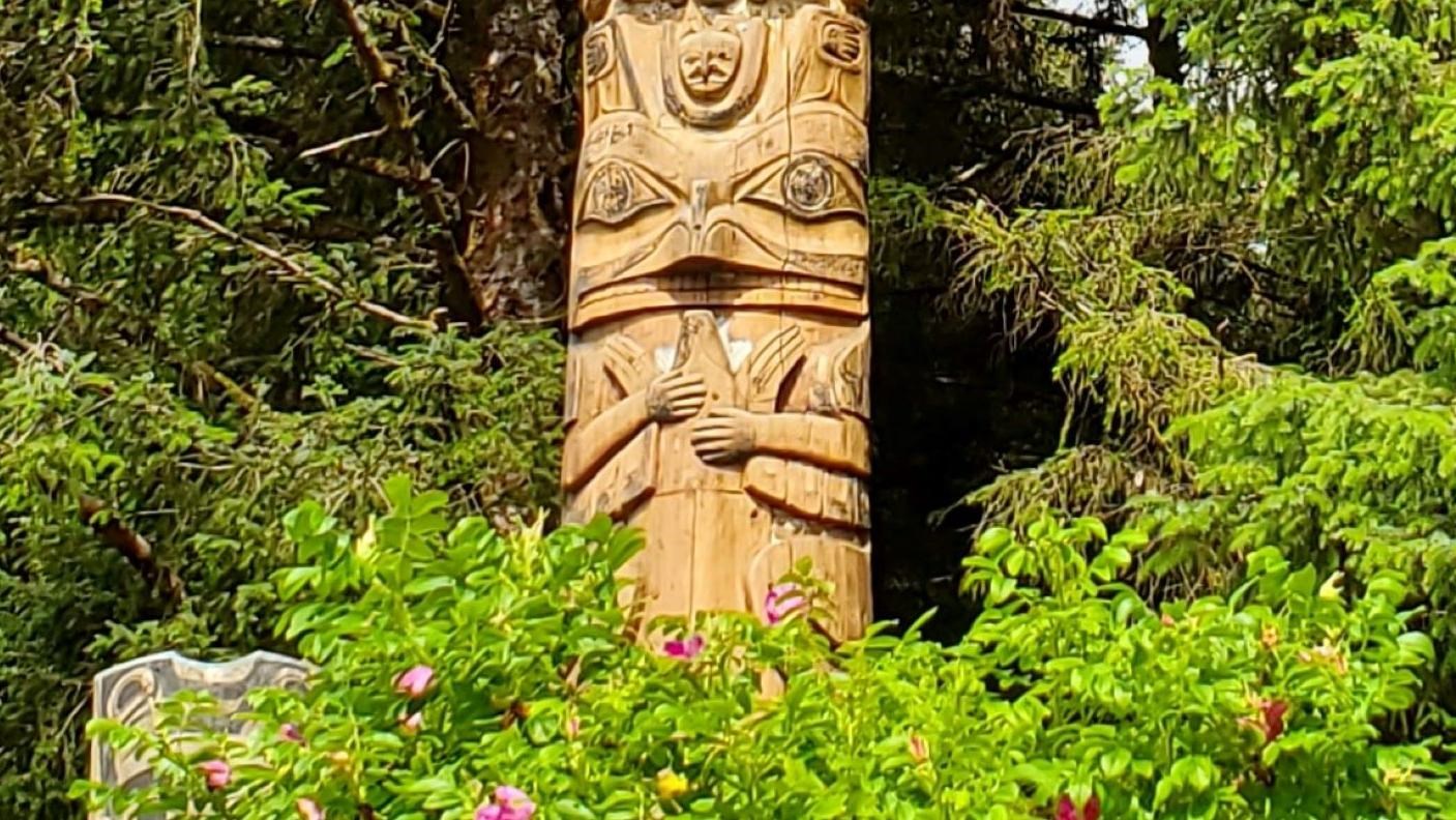 A portion of a large totem pole behind a rose bush with bright pink flowers and in front of a forest