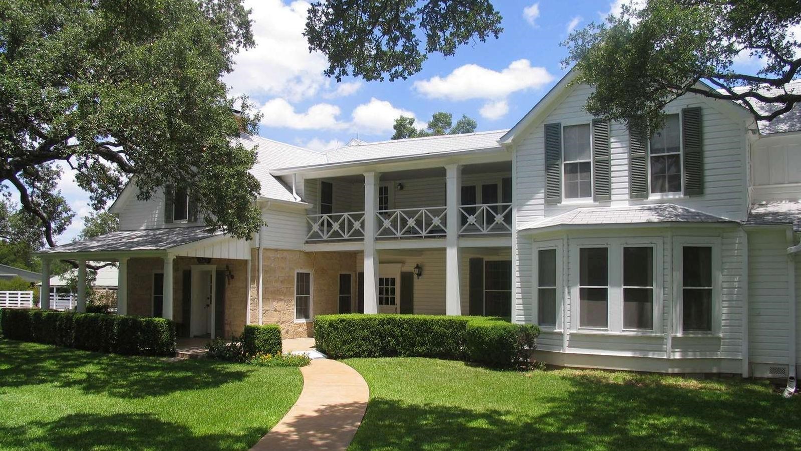 Shade from trees covers a green lawn and a sidewalk leads up to a large, white, two-story home.