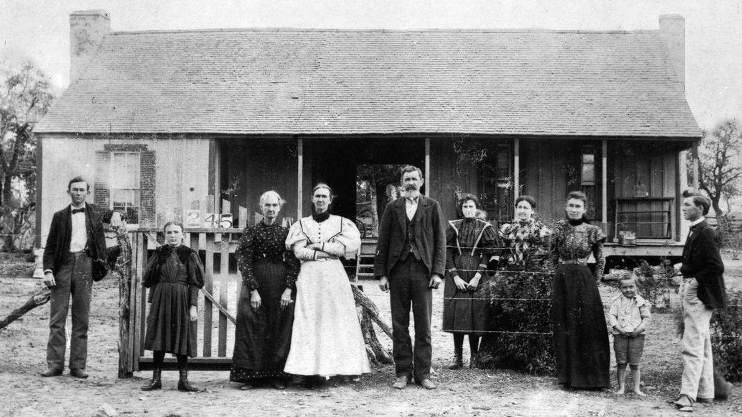 A well dressed family of ten stand in front of a worn, one story ranch house on a barren landscape.