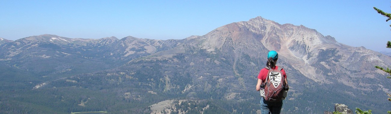 A woman wearing a backpack stands on the top of a mound of rocks and looks out across the valley