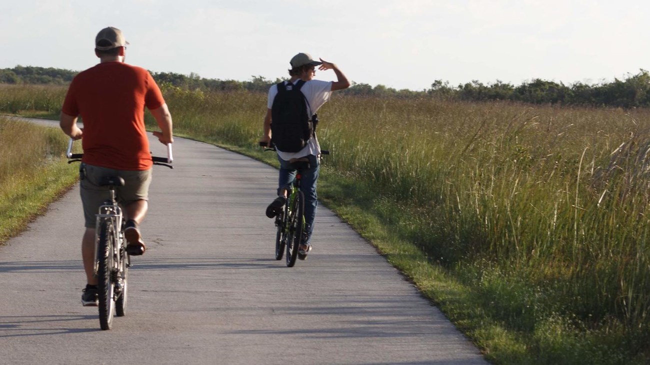 Two bikers peddle along a paved road flanked by open prairie. One bikers stares over the horizon