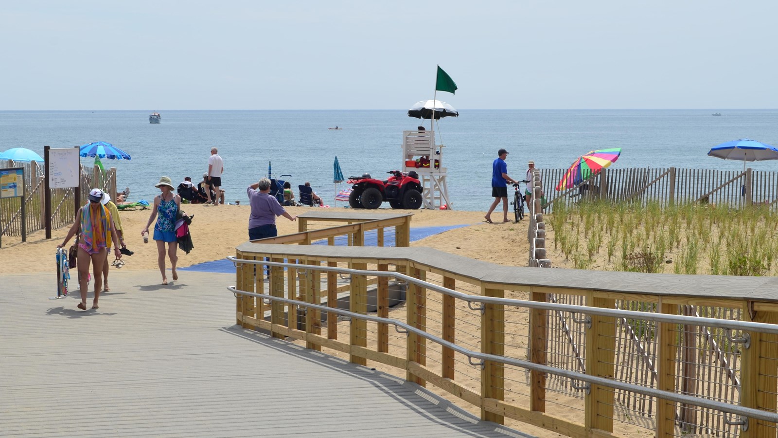 People walk on a wide walkway with handrail leads down to a sandy beach.