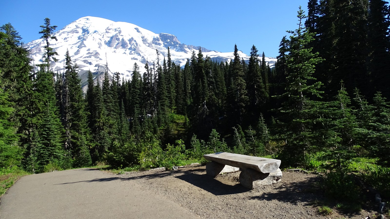 A wide paved trail with a low log bench to the right. Mount Rainier rises above the trail.