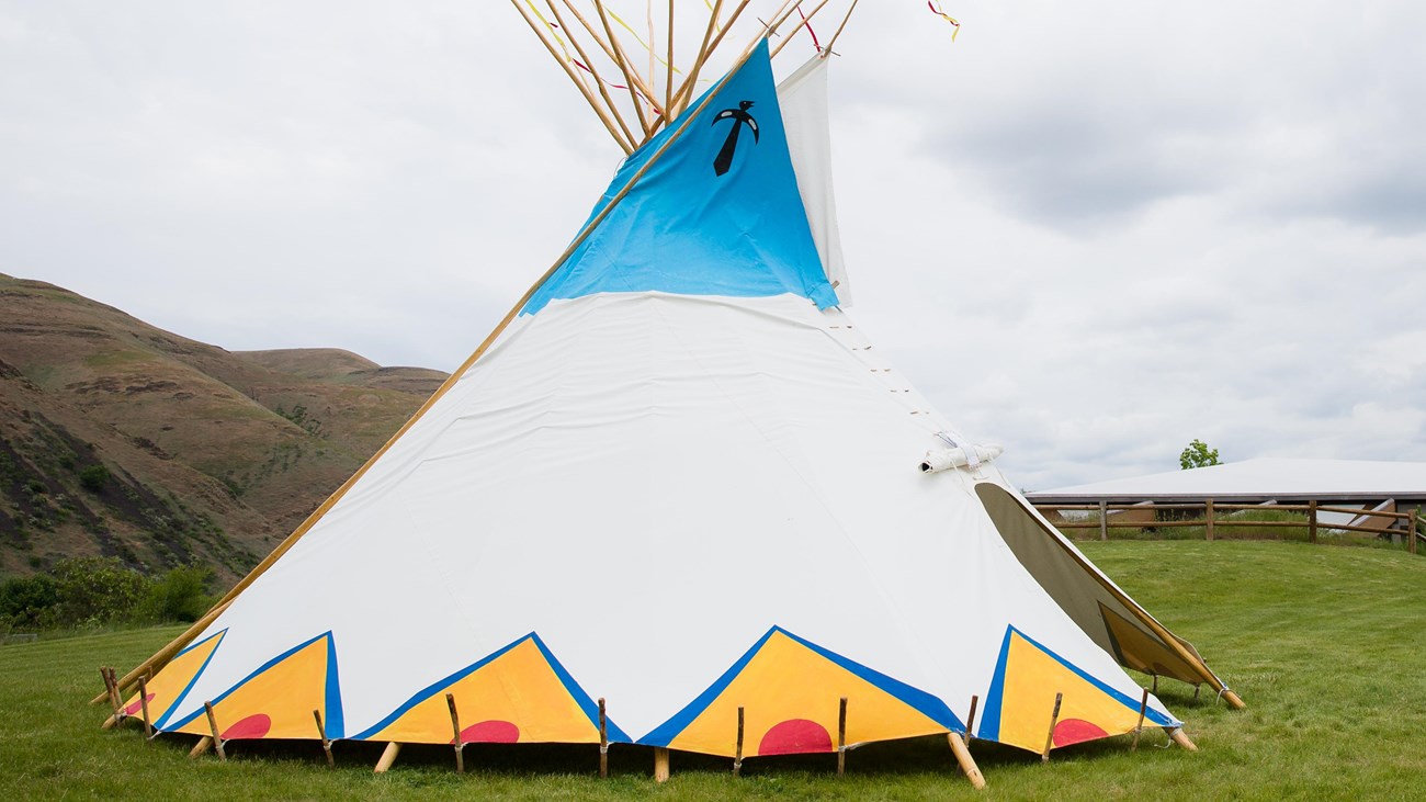 Color photograph of a tipi pitched on the grass in front of a low-slung building.