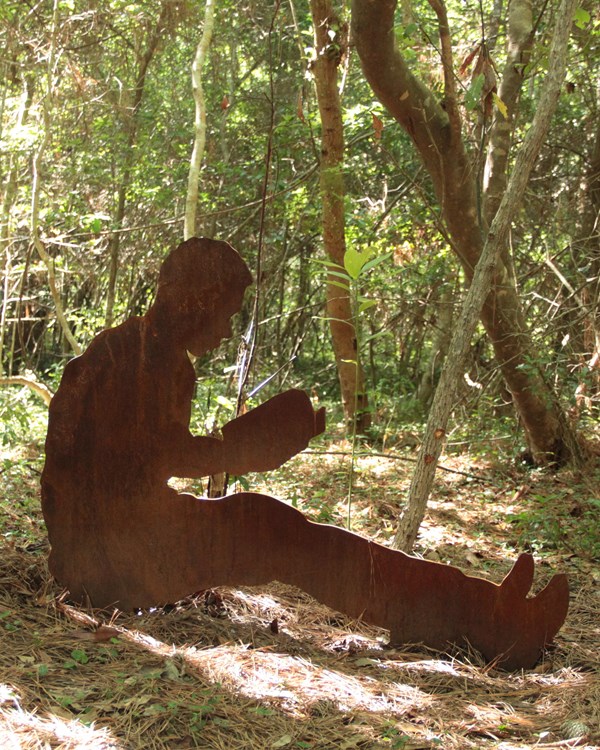 A metal silhouette of a boy learning against a tree reading. Behind him is forested area