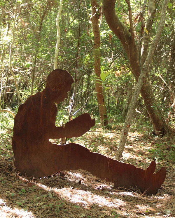 A metal silhouette of a boy learning against a tree reading. Behind him is forested area