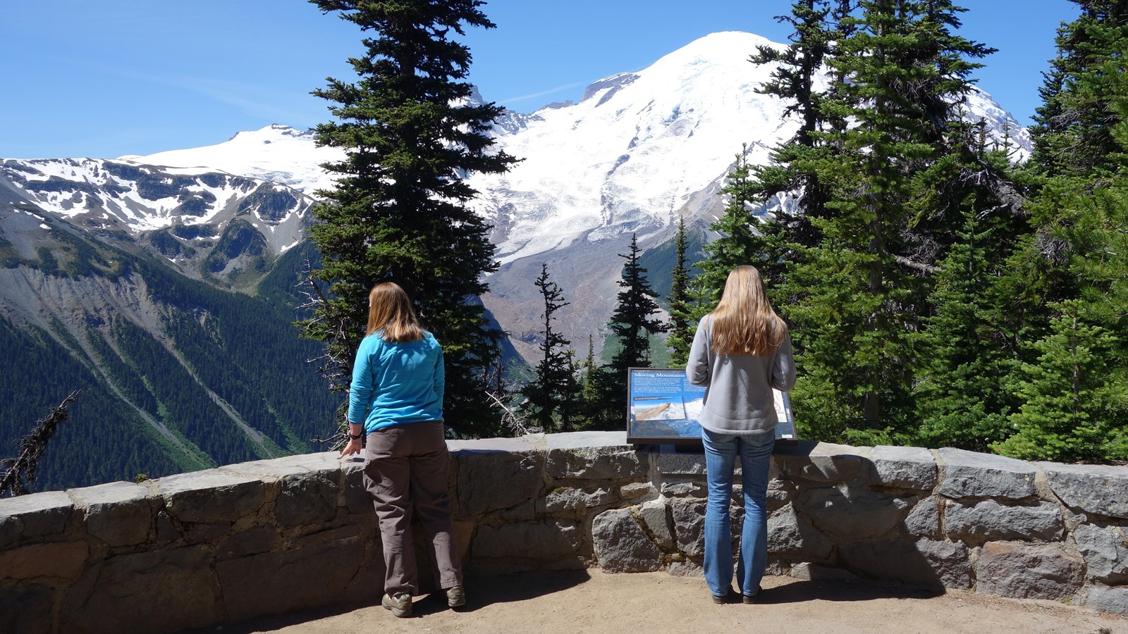 Two women stand next to a low rock wall looking at a view of a glaciated mountain and river valley.