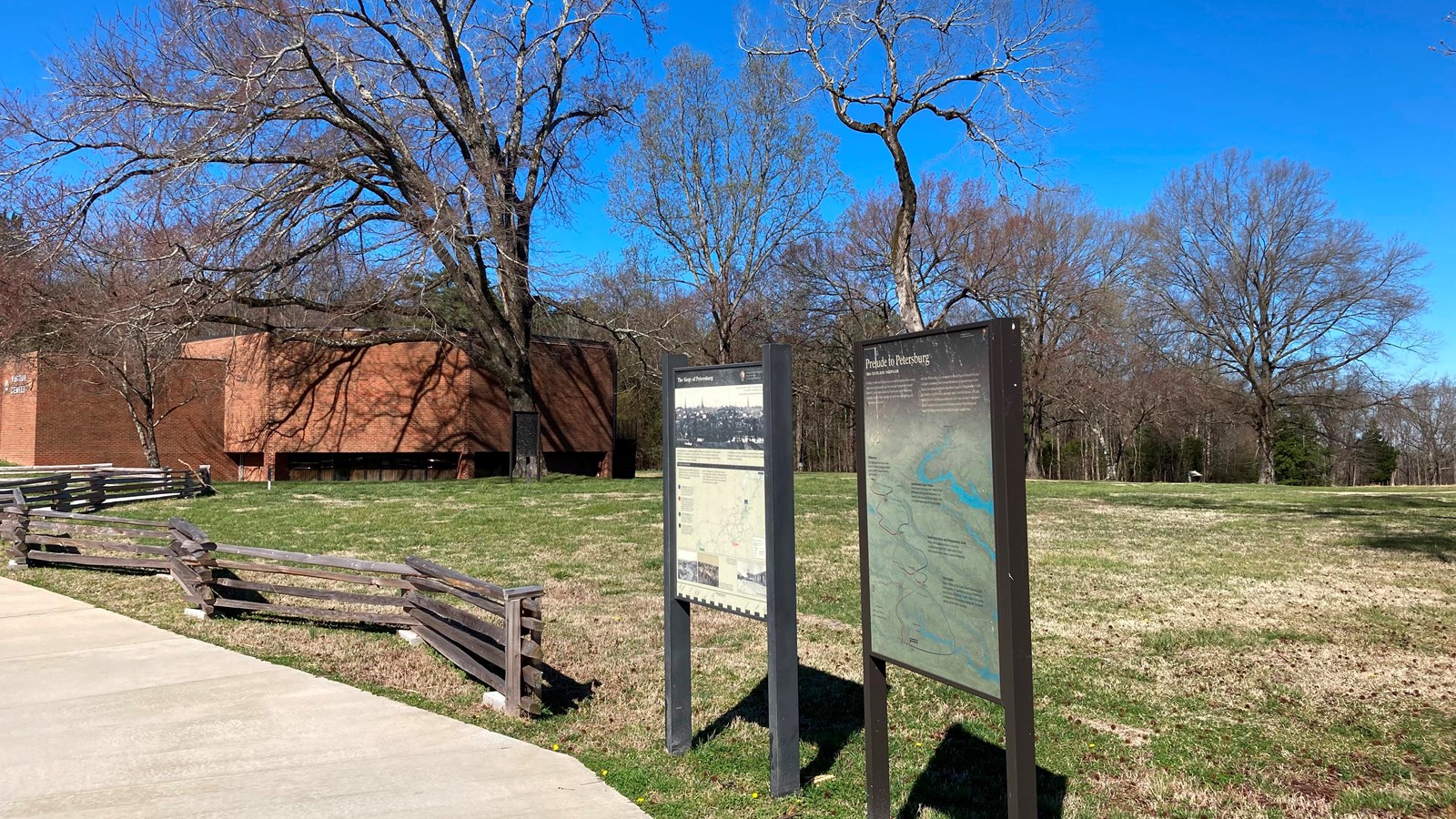 Two vertical signs next to sidewalk and low split rail fence. Red brick building in background.