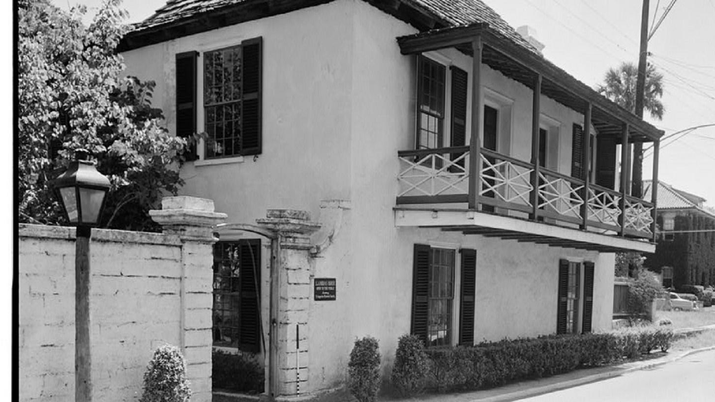 White two story house with a second story balcony and dark roof shudders and a wall to the left