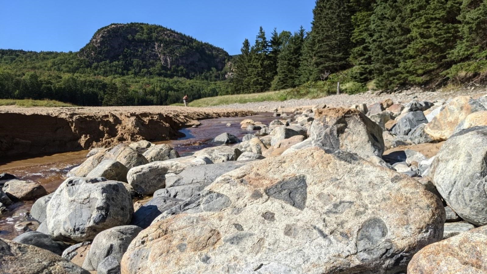 A boulder with chunks of rock inside it sits in front of a beach and mountain.