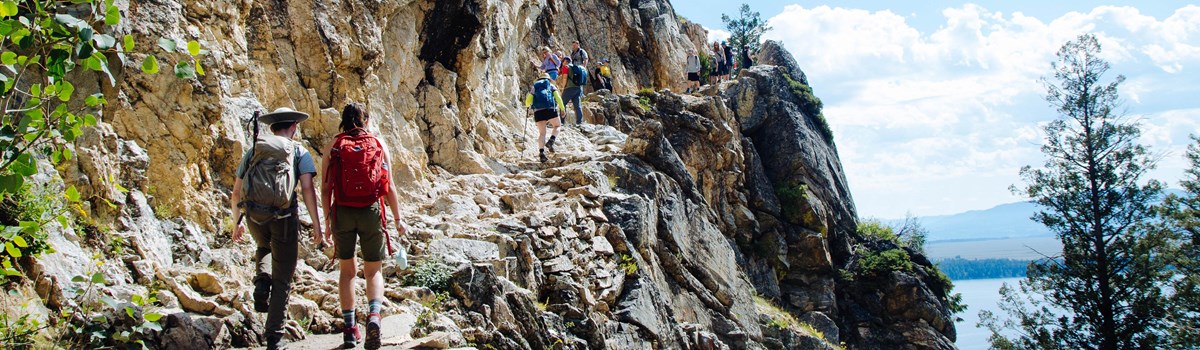 Hikers walk up a rocky trail with a cliff on the right.