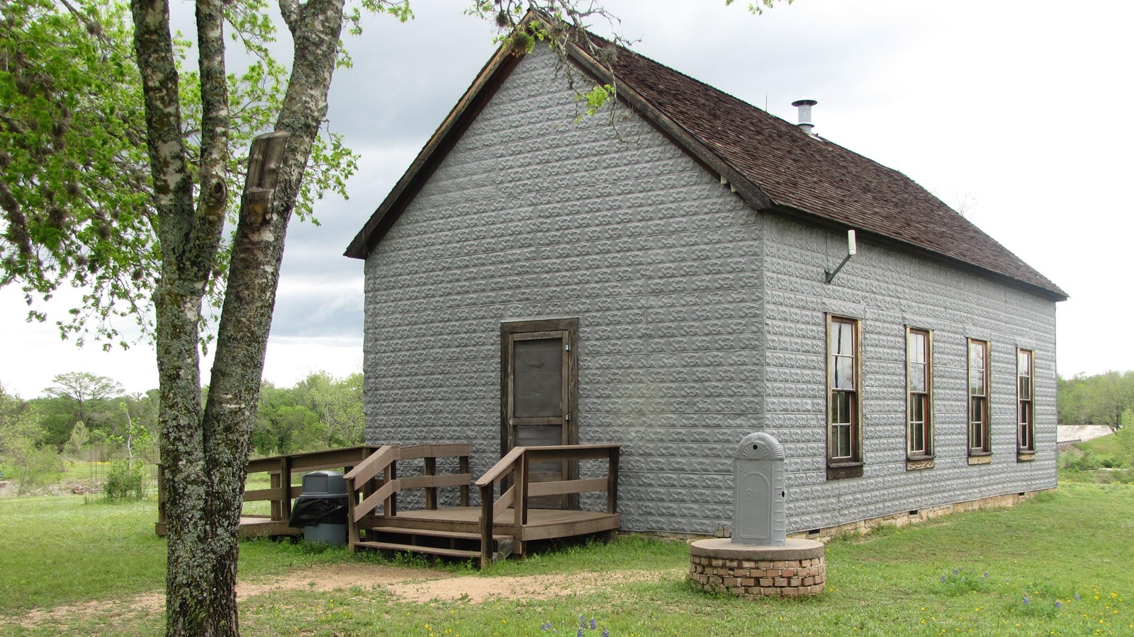 A gray building with a shingled roof has tall windows on its side and a stepped entrance at its end.