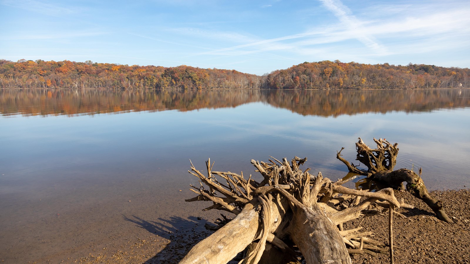 A gravel lot edged by a small wooden fence borders a river. A bright blue sky shines on the scene. 