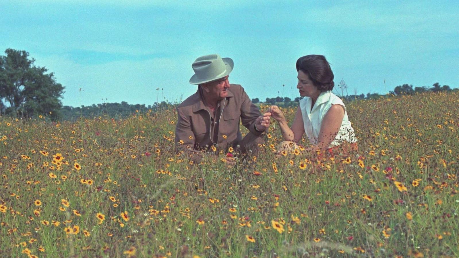 President and Mrs. Johnson touch hands while sitting in a large field of yellow wildflowers.