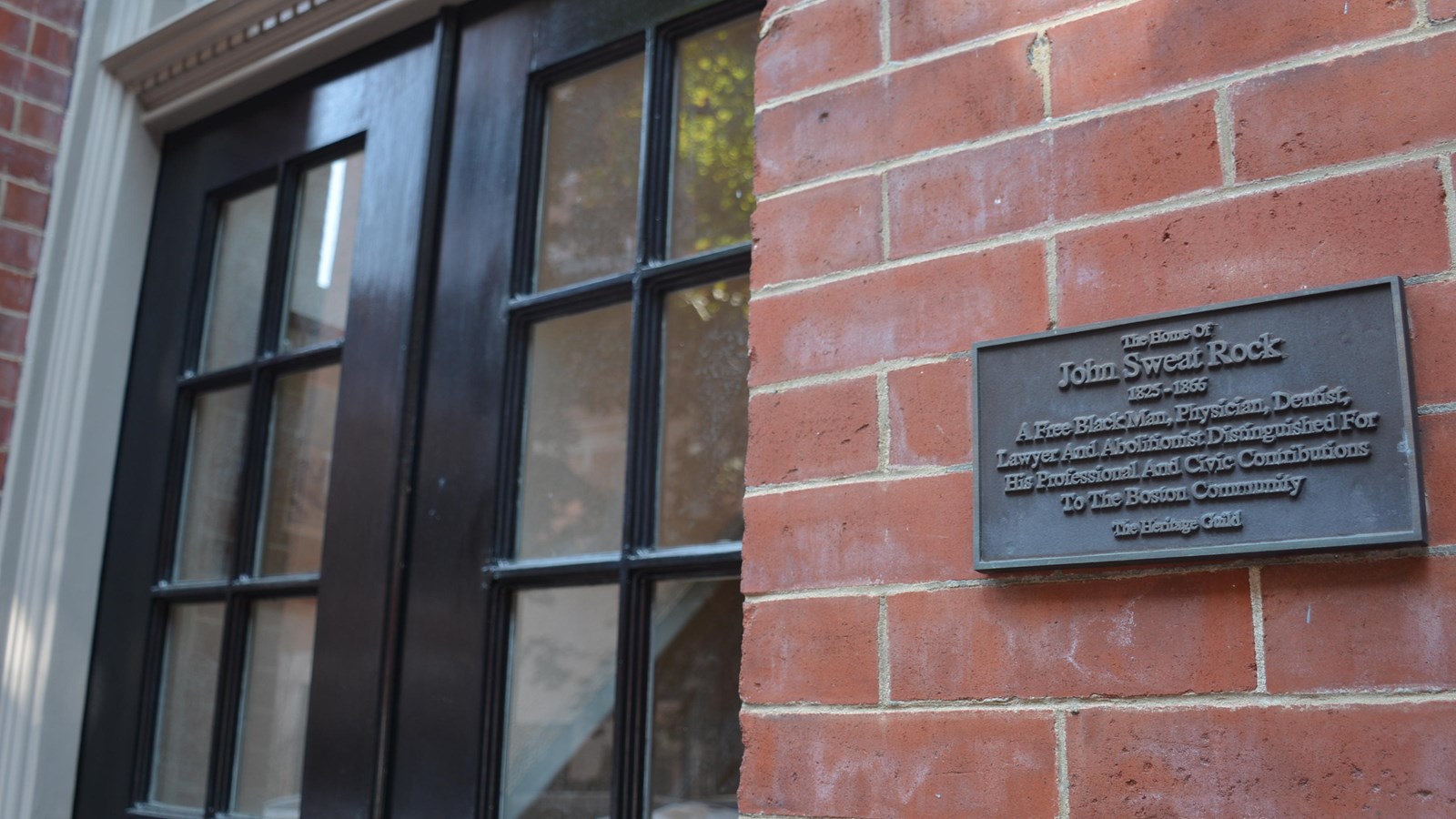 Close-up of a brick building\'s entrance. The door has window panes and there is a historic plaque.