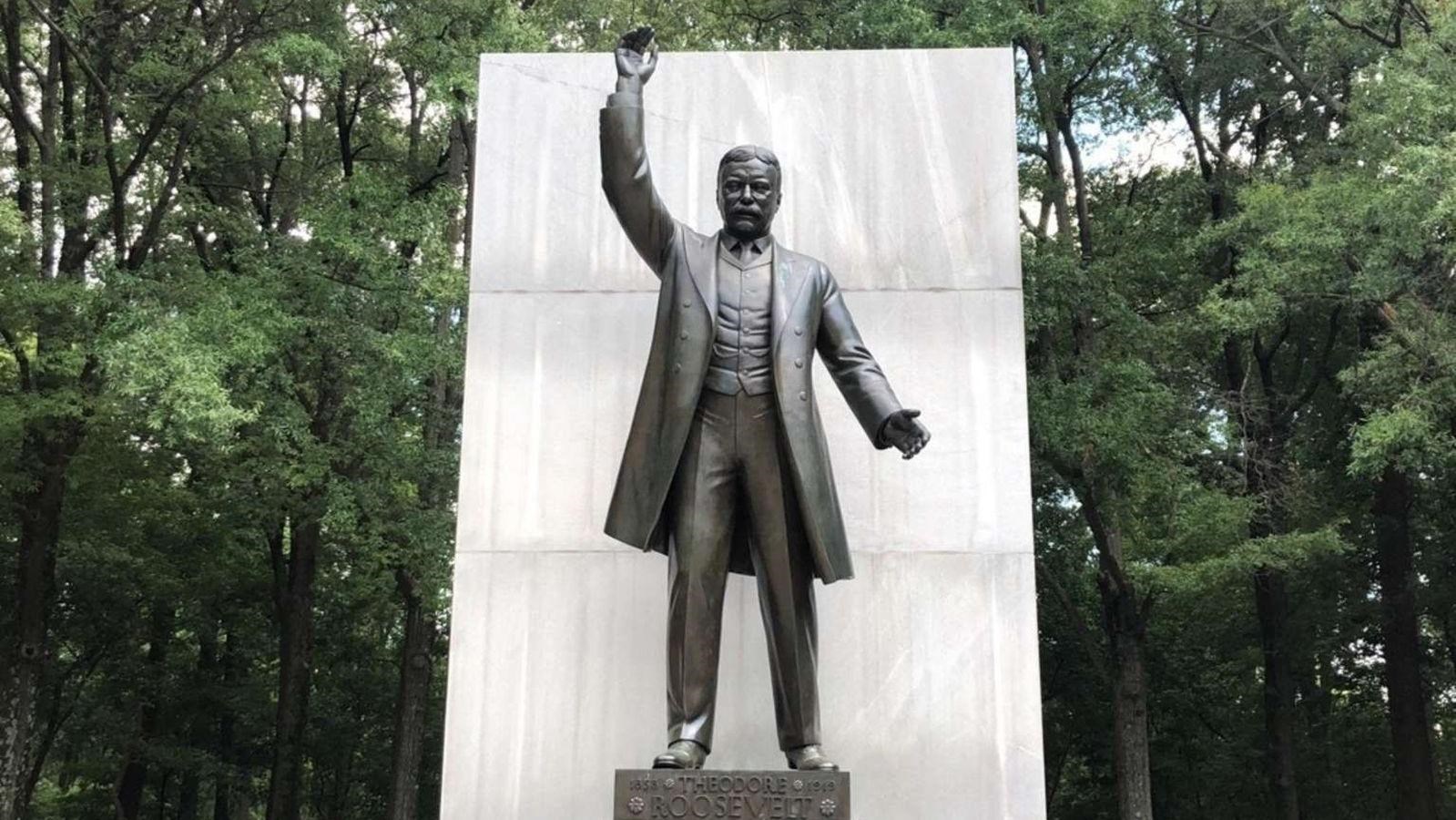 A tall bronze statue of Theodore Roosevelt stands in front of several trees.