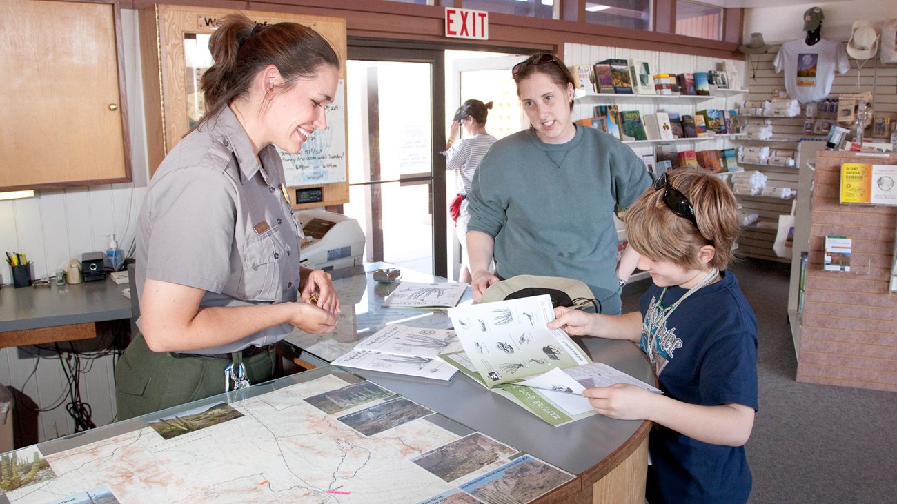 A female ranger reviews a young boy\'s junior ranger booklet, with mom watching from the side.